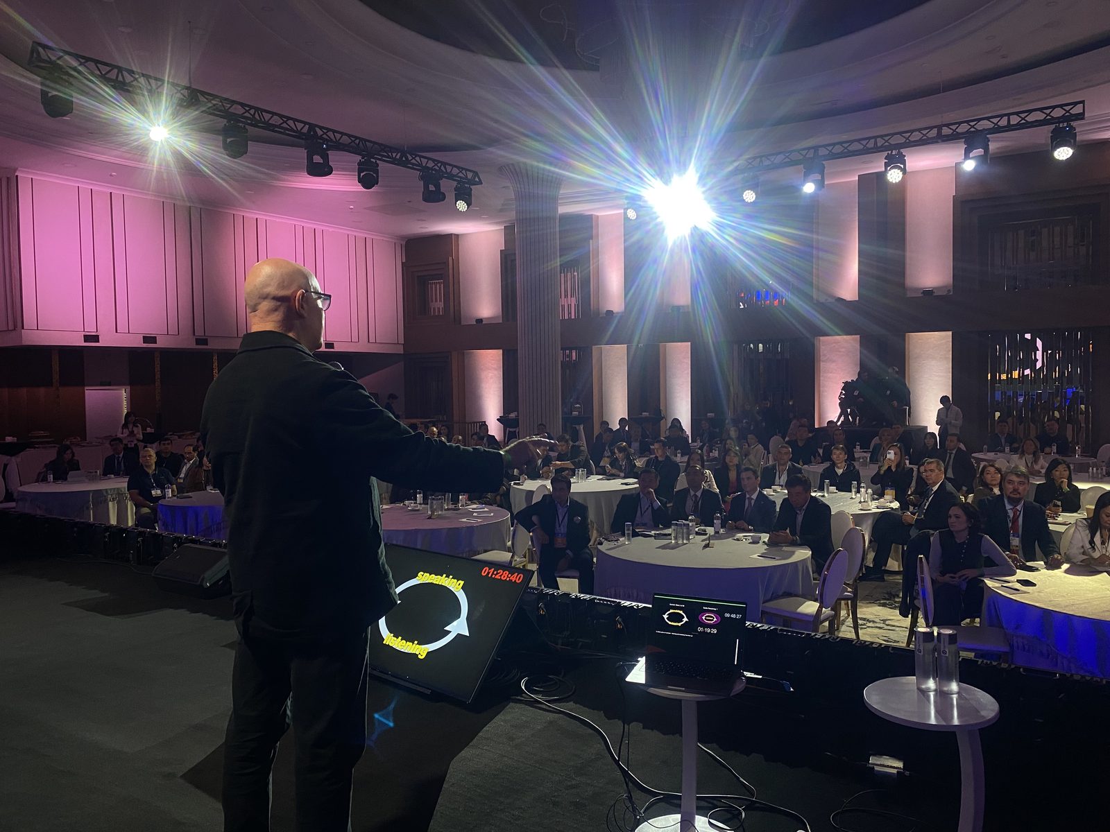 Julian Treasure, seen from behind on stage, gesturing to a ballroom audience at round tables under bright stage lights