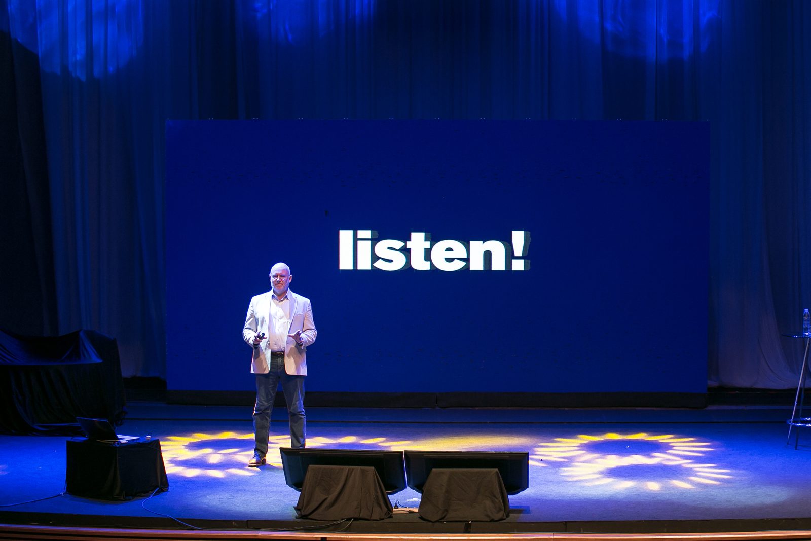 Julian Treasure in a light jacket on a blue-lit stage in front of a large screen reading listen!, Bangkok