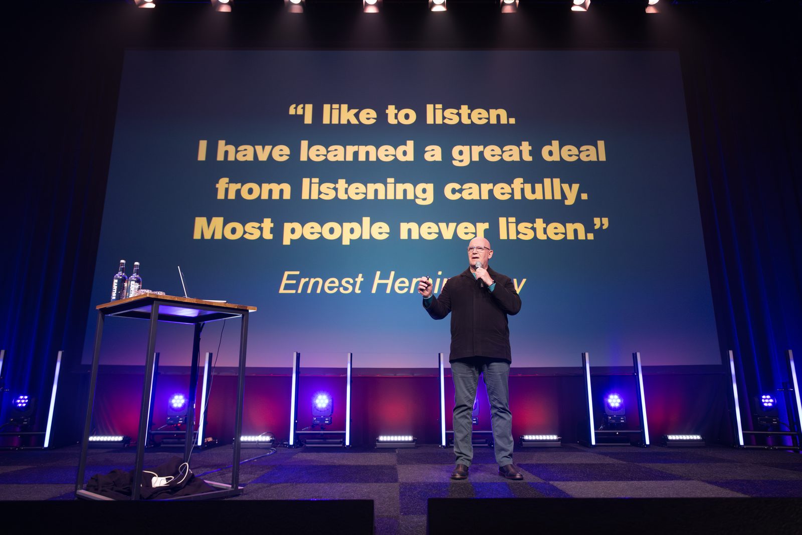 Julian Treasure speaking into a handheld microphone in front of a screen quoting Ernest Hemingway on listening, Rotterdam